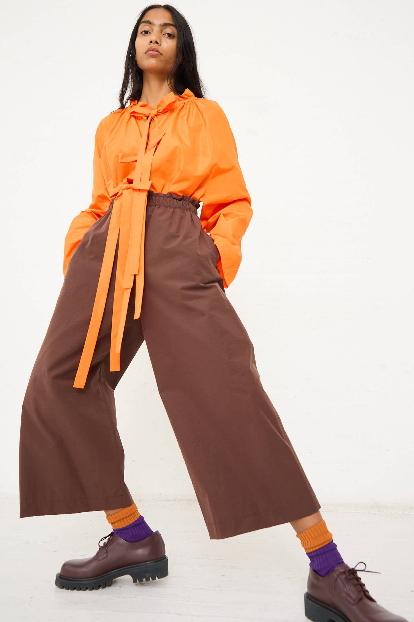 Person wearing an orange long-sleeve top with a tie, AVN's Easy Pant in brown with a ruffled waist, orange and purple socks, and brown shoes. They are standing against a plain white background.