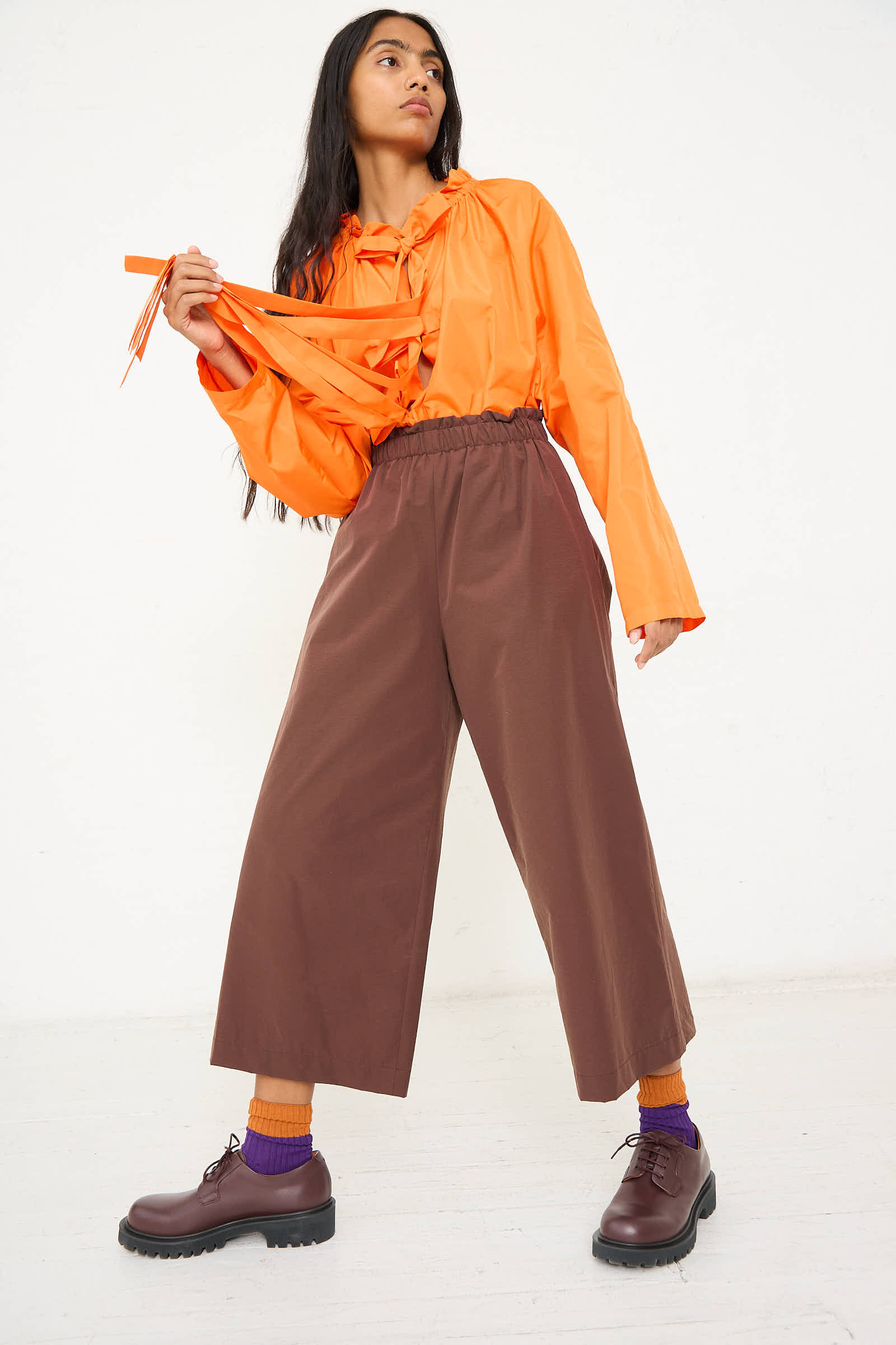 A person in an orange long-sleeve shirt and the Easy Pant in Brown by AVN stands against a plain background, holding orange straps. They are wearing brown shoes and purple socks.