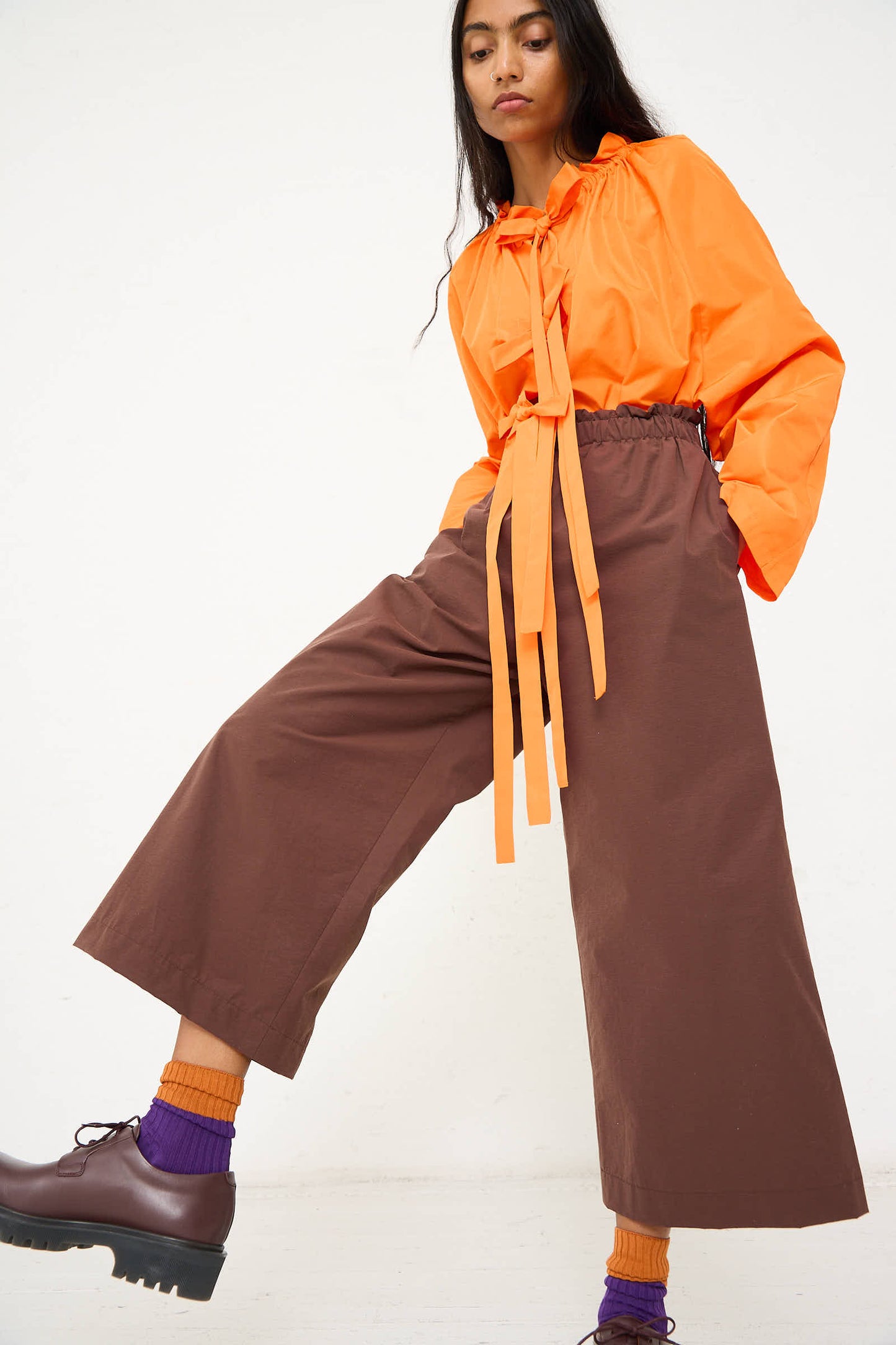 Person wearing an orange long-sleeve top with a tie detail, AVN's Easy Pant in Brown, purple socks, and brown platform shoes, posing against a plain white background.