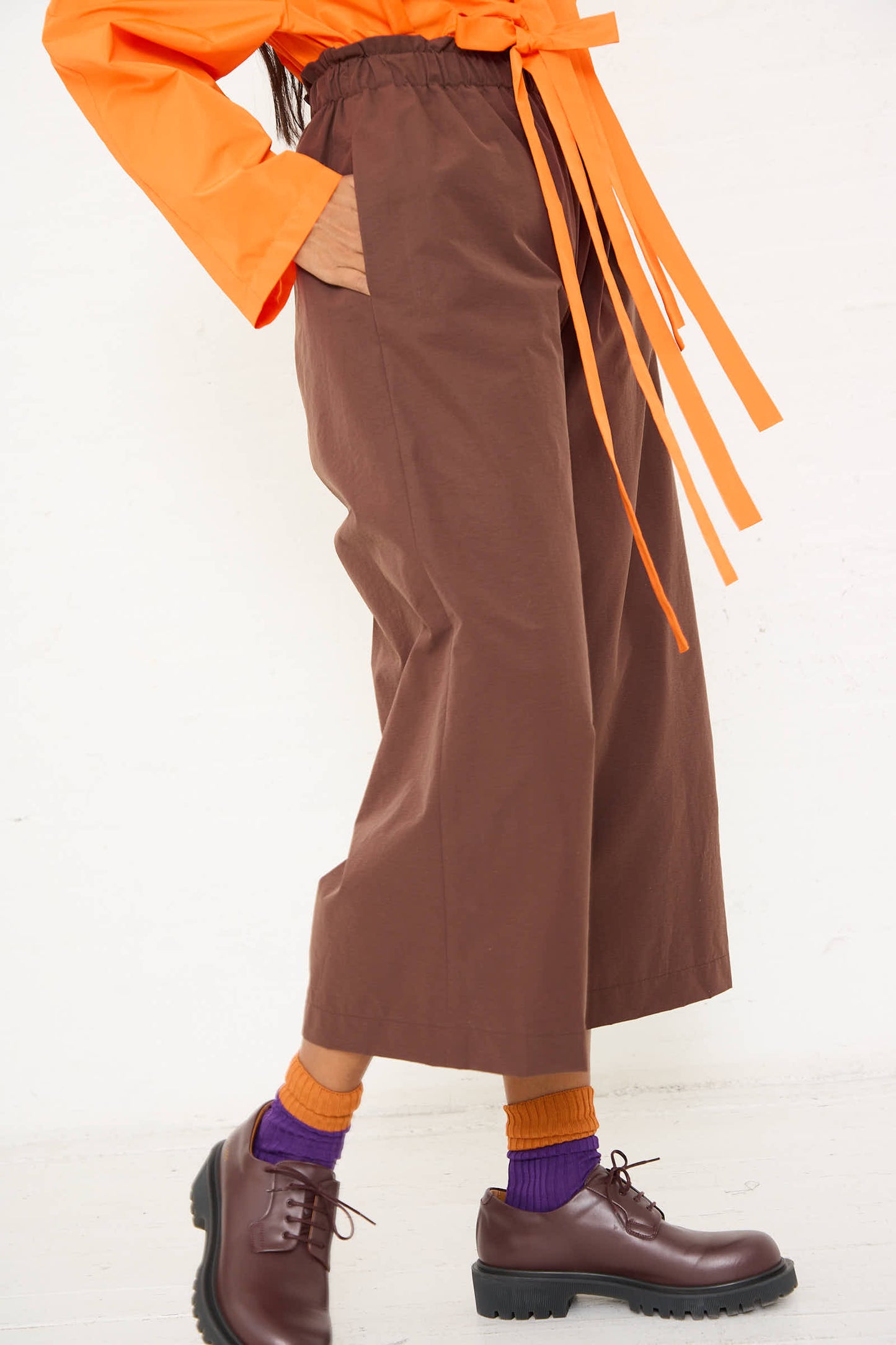 A person wearing AVN's Easy Pant in Brown with a ruffled waist, an orange top, and dark brown shoes with purple and orange socks, standing against a light background.