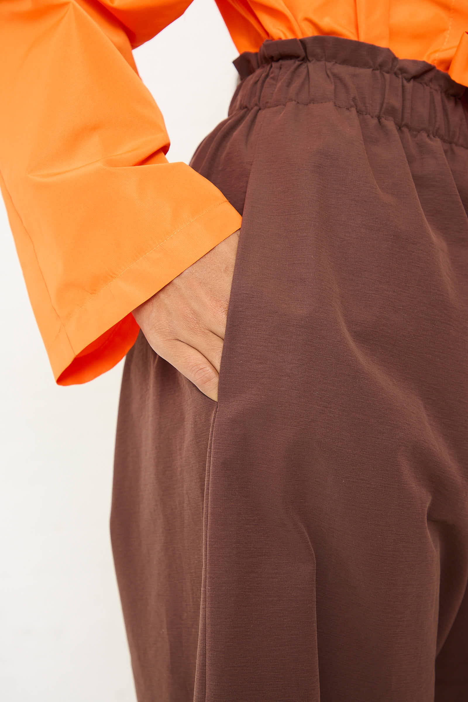 Close-up of a person wearing an AVN Easy Pant in Brown, featuring wide-leg design and an elastic waistband. They're also wearing an orange long-sleeve top, with their hand casually placed in their pocket.