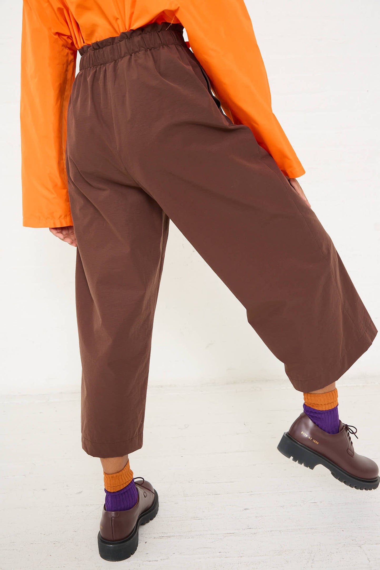 A person is shown from the back wearing a bright orange shirt, the AVN Easy Pant in Brown with a ruffled waist, orange socks, purple socks layered over the orange ones, and brown shoes.