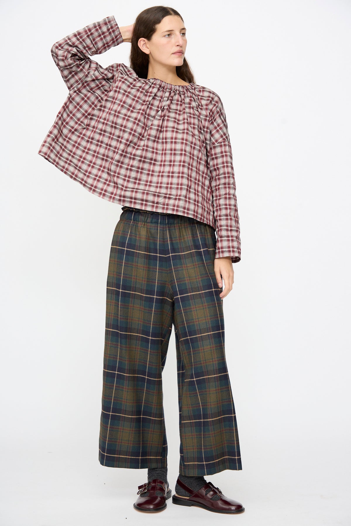 A person wearing a long-sleeved, burgundy plaid blouse and AVN Easy Pants in Military Check, styled with dark burgundy buckle shoes.
