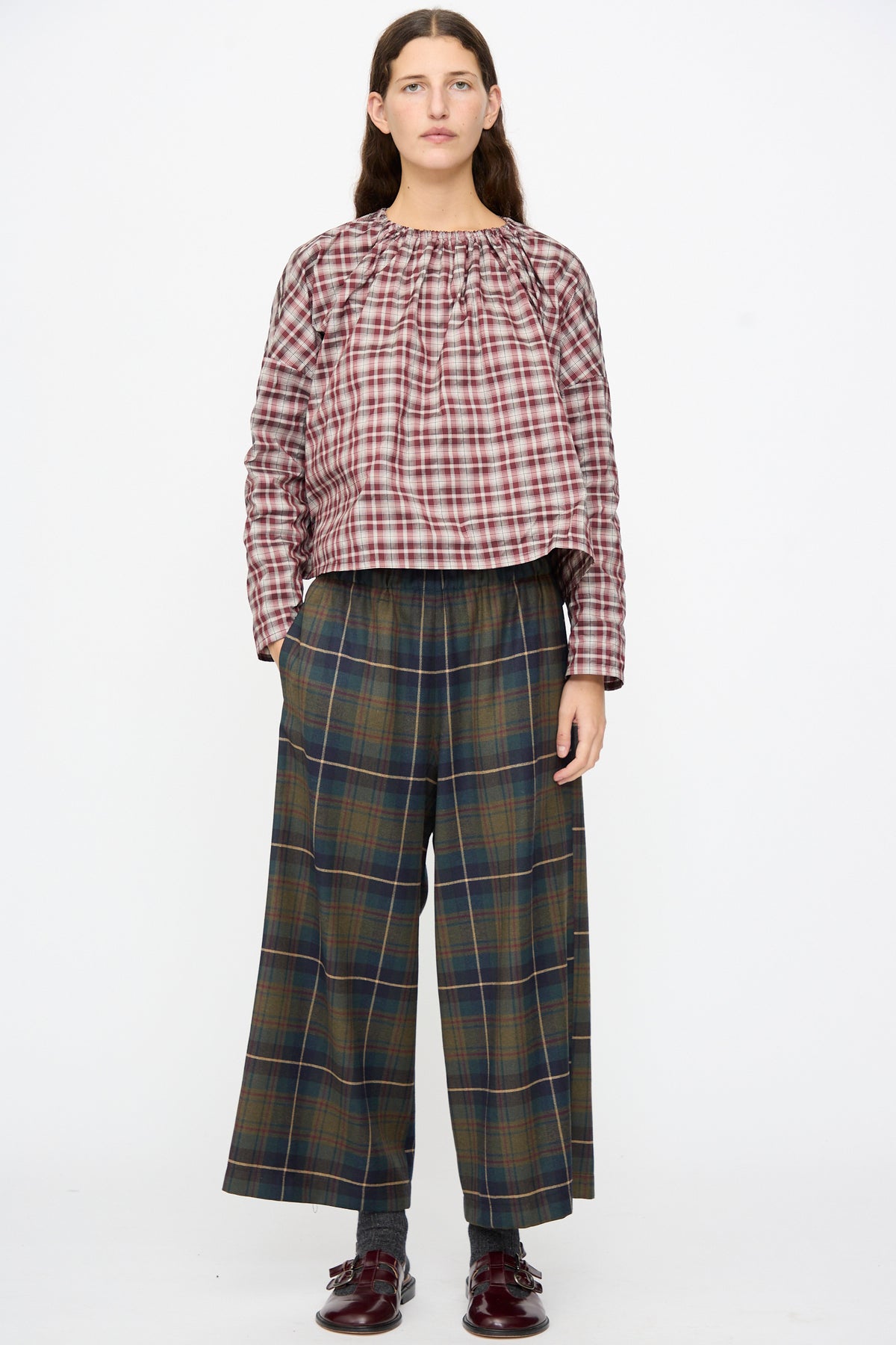 A person wears a loose-fitting, red-and-white plaid long-sleeve top and AVN Easy Pants in Military Check, featuring a wide-leg, dark green plaid pattern.