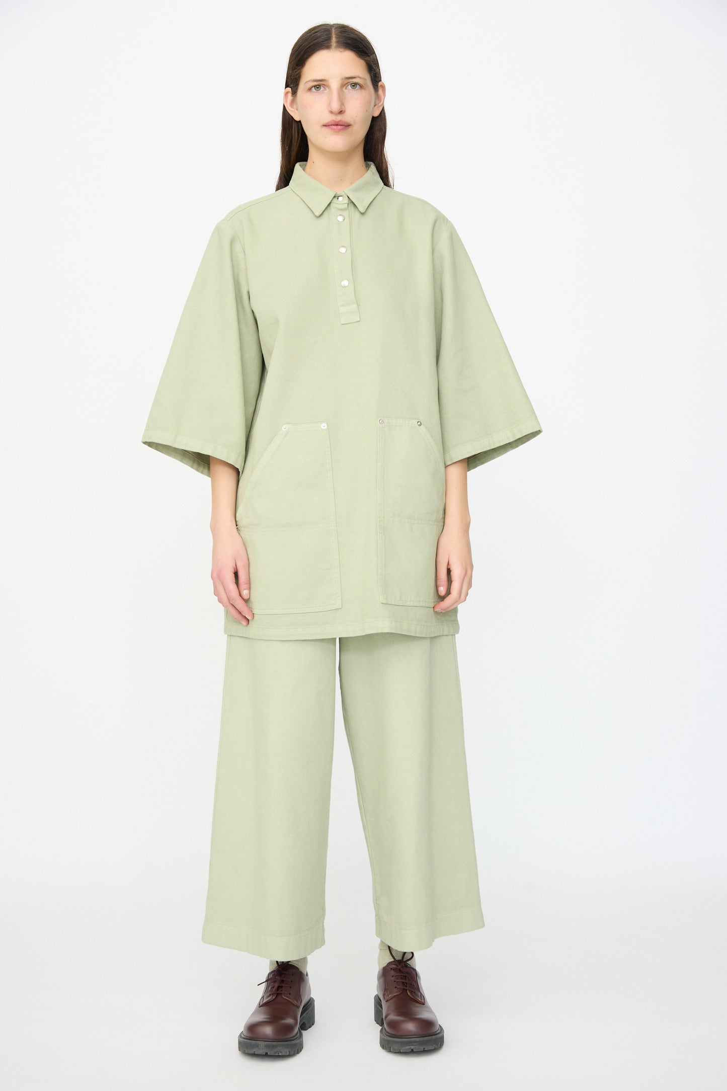 A person stands facing forward wearing AVN’s Easy Pants in Sage, a loose wide-leg cotton style made in Italy, paired with a light green collared top with large front pockets and dark brown shoes.