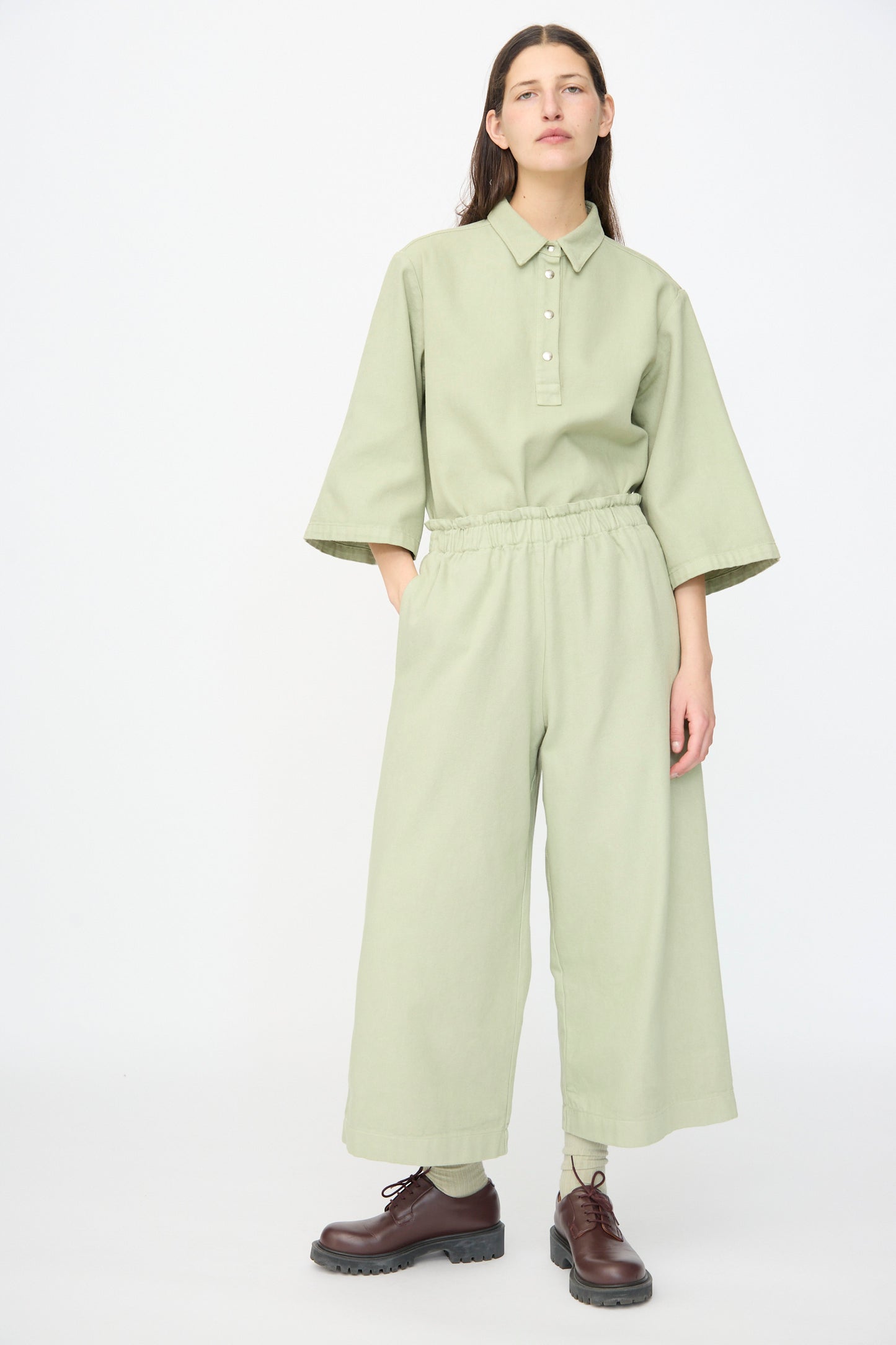 A person stands against a plain background wearing AVN’s Easy Pants in Sage, paired with a matching sage green collared top and dark brown lace-up shoes. The wide-leg cotton pants are made in Italy.