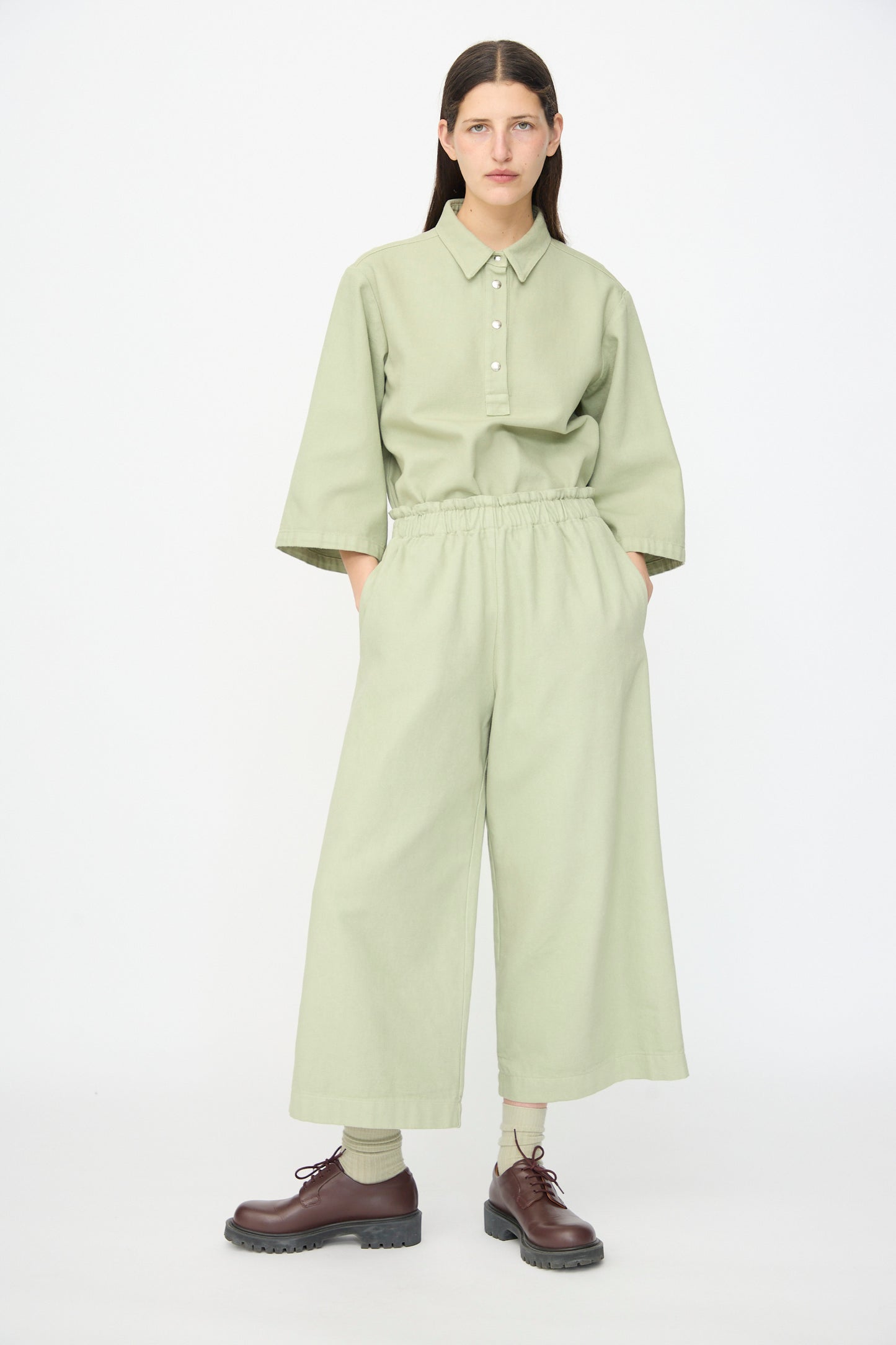 A person stands facing forward wearing a light green button-up shirt, AVN Easy Pants in Sage (made in Italy), and brown lace-up shoes.