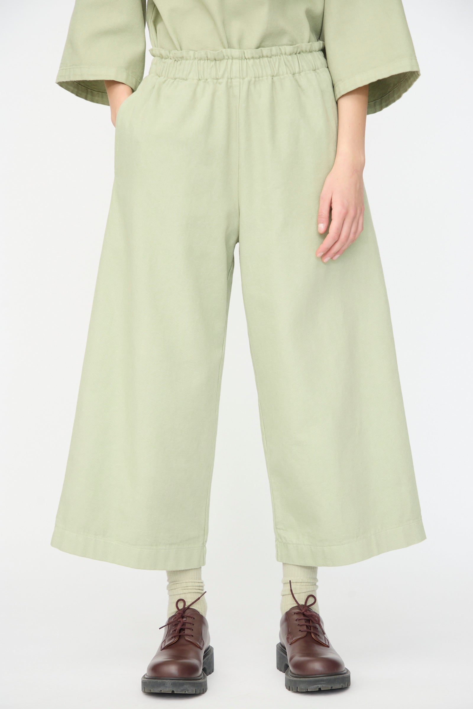 A person wearing AVN’s Easy Pants in Sage with a matching top, beige socks, and brown lace-up shoes stands against a plain white background.