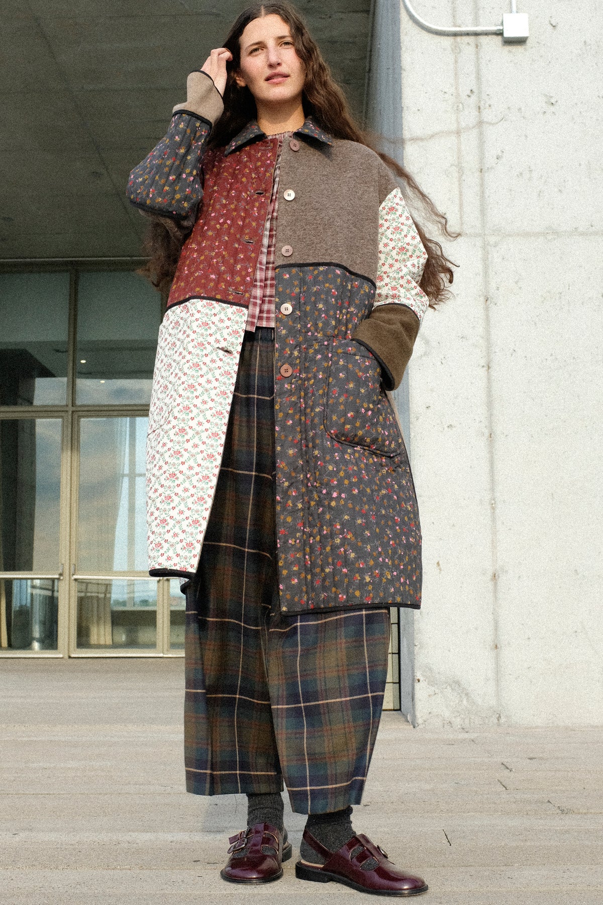 A woman standing outside a building wearing AVN's Mid Season Coat in Camel, Bolla Shirt in Bordeaux Check. and Easy Pants in Military Check.