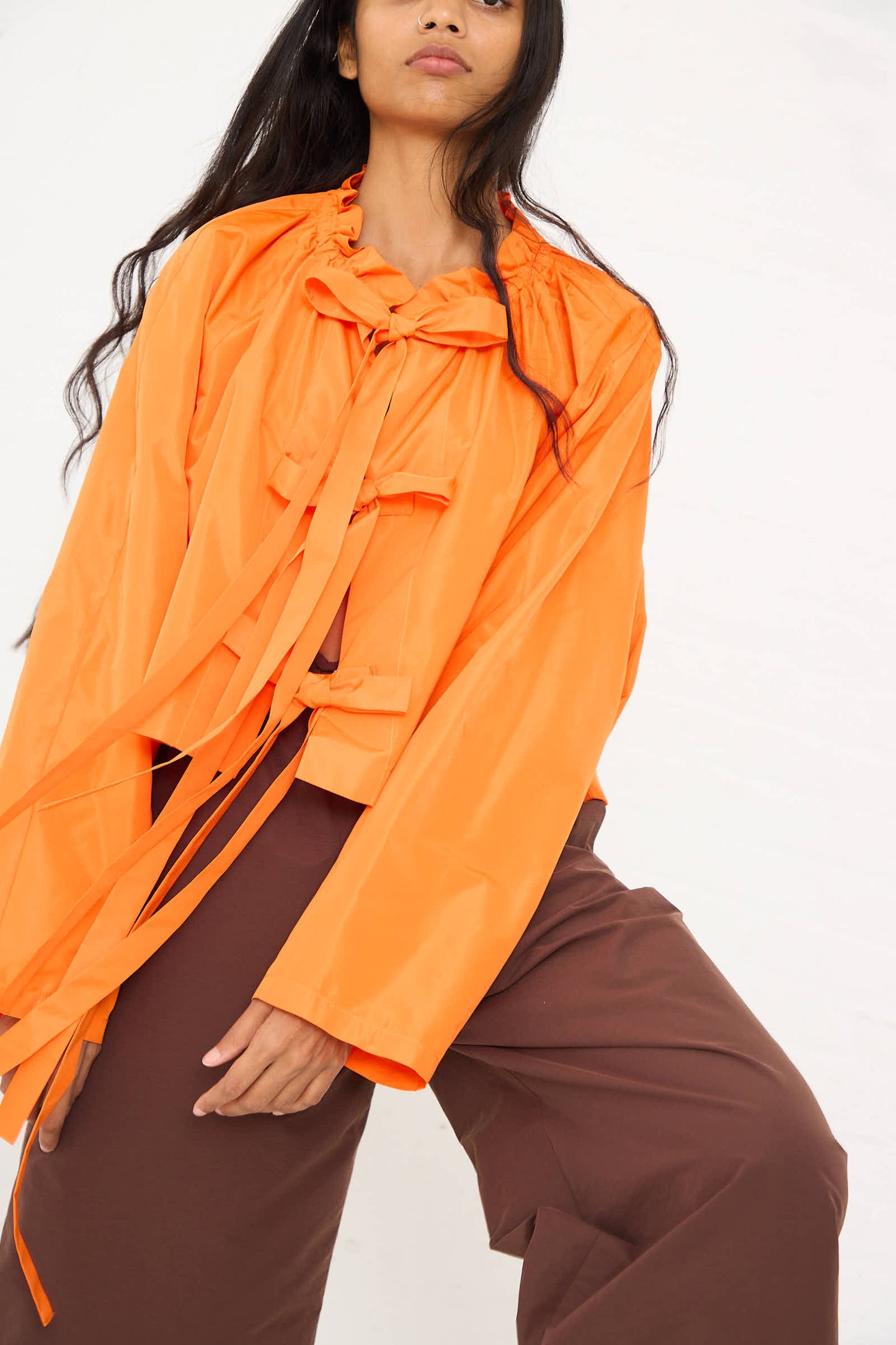 Person wearing the AVN "Frappa Shirt" in bright orange taffeta, featuring long sleeves, a ruffled neck, and long ribbons, paired with brown pants and posing against a plain background.