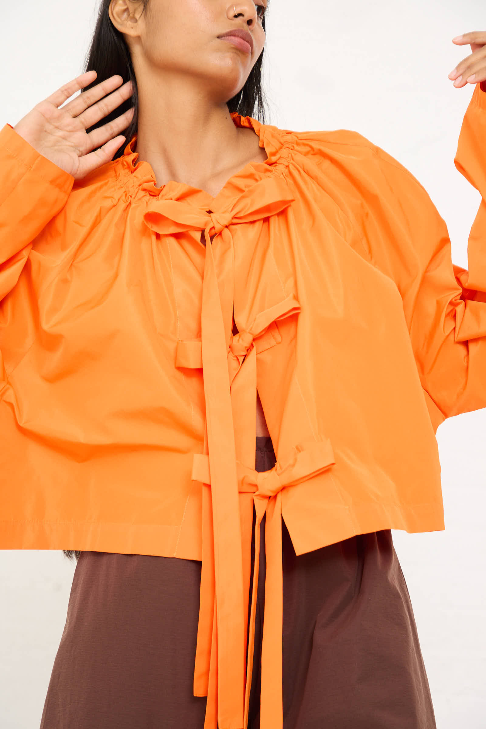 Person wearing the AVN Frappa Shirt in bright orange taffeta, featuring long sleeves with ribbon ties and paired with brown pants, posing with arms raised and hands near face. The relaxed fit and ruffled neck add an elegant touch to the look.