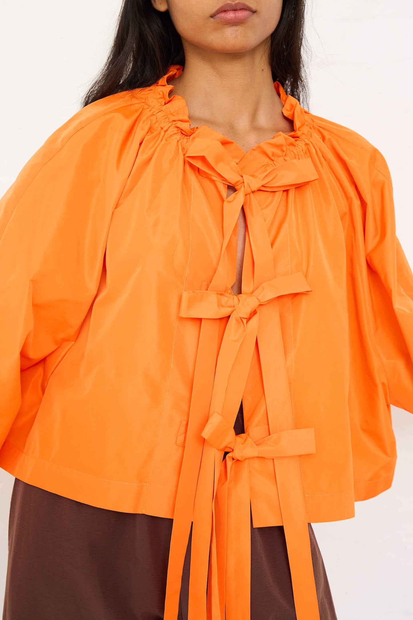 A person wearing the AVN Frappa Shirt in Orange, a relaxed-fit, bright orange taffeta top that features multiple tied bows on the front and a ruffled neck.