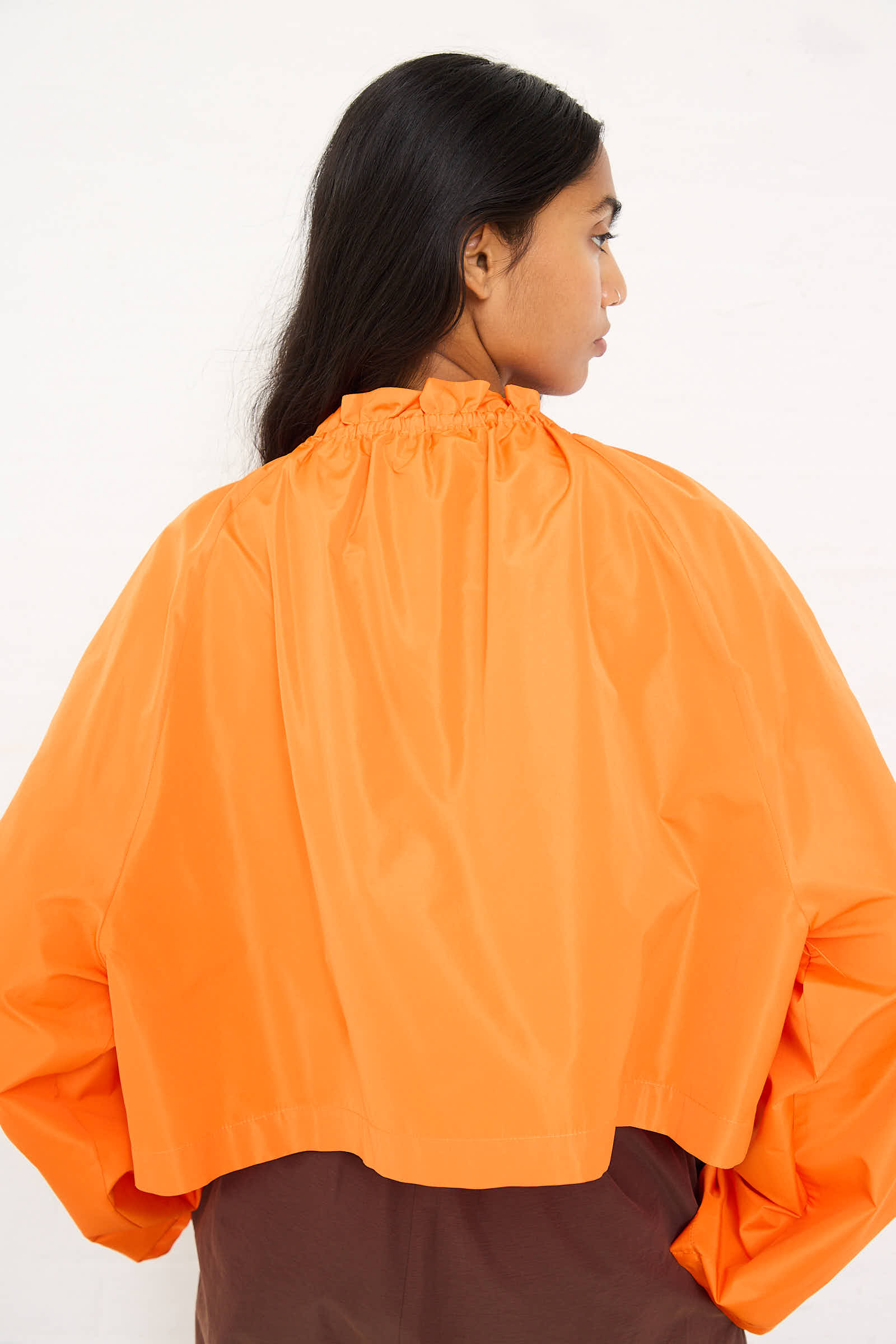 Person standing with back facing camera, wearing an AVN Frappa Shirt in Orange featuring long sleeves, a relaxed fit, and a ruffled neckline, paired with brown bottoms, all set against a plain white background.