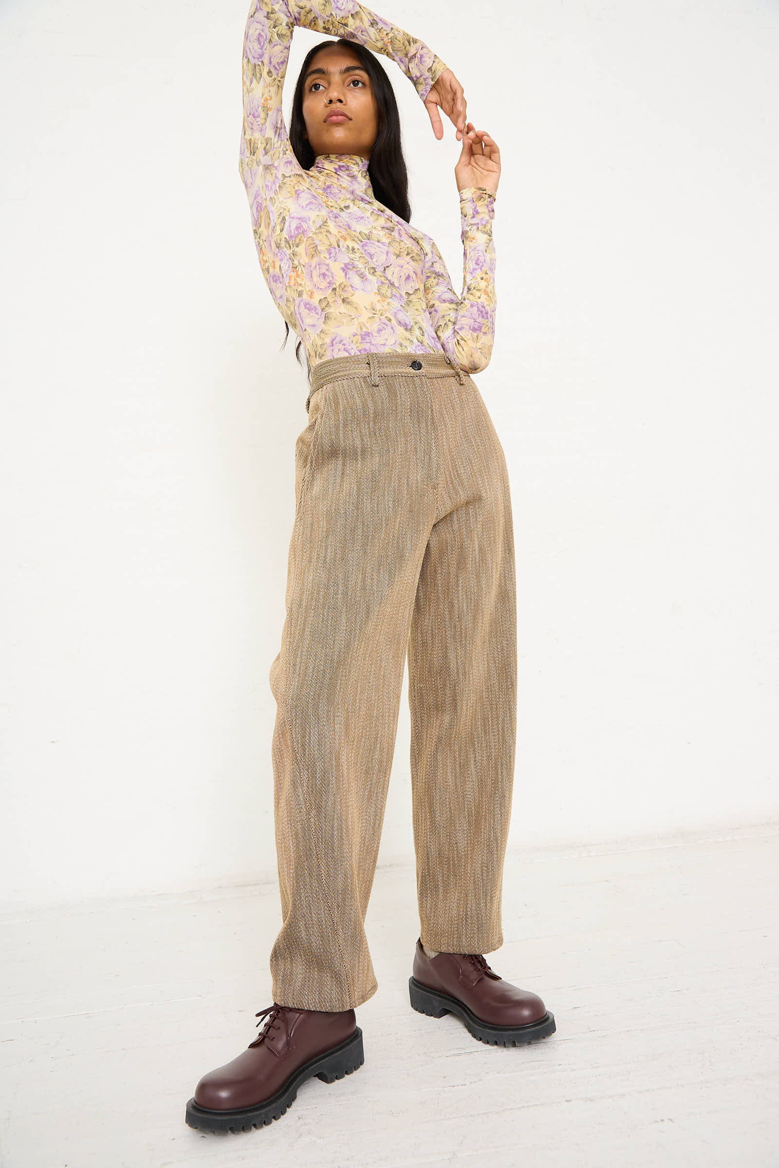 A woman stands against a white background, wearing a floral long-sleeve top, the AVN Iris Pant in Beige with side slash pockets, and brown boots. Her left arm is bent with her hand resting on her head, while her right arm is raised. Made in Italy.