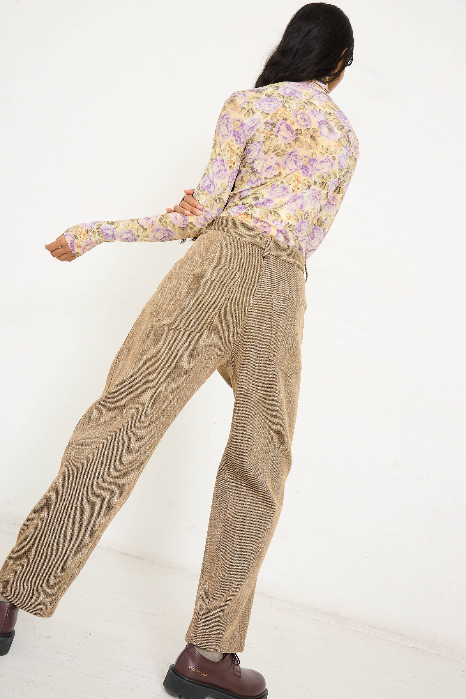 A person wearing a floral long-sleeve top, the Iris Pant in Beige by AVN with side slash pockets, and brown shoes is standing with their back to the camera against a plain white background.