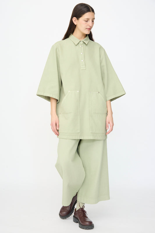 Someone wearing the AVN Joyeux Dress in Sage, a loose-fitting, textured cotton dress with front pockets and a collar, stands against a plain background, styled with brown lace-up boots.