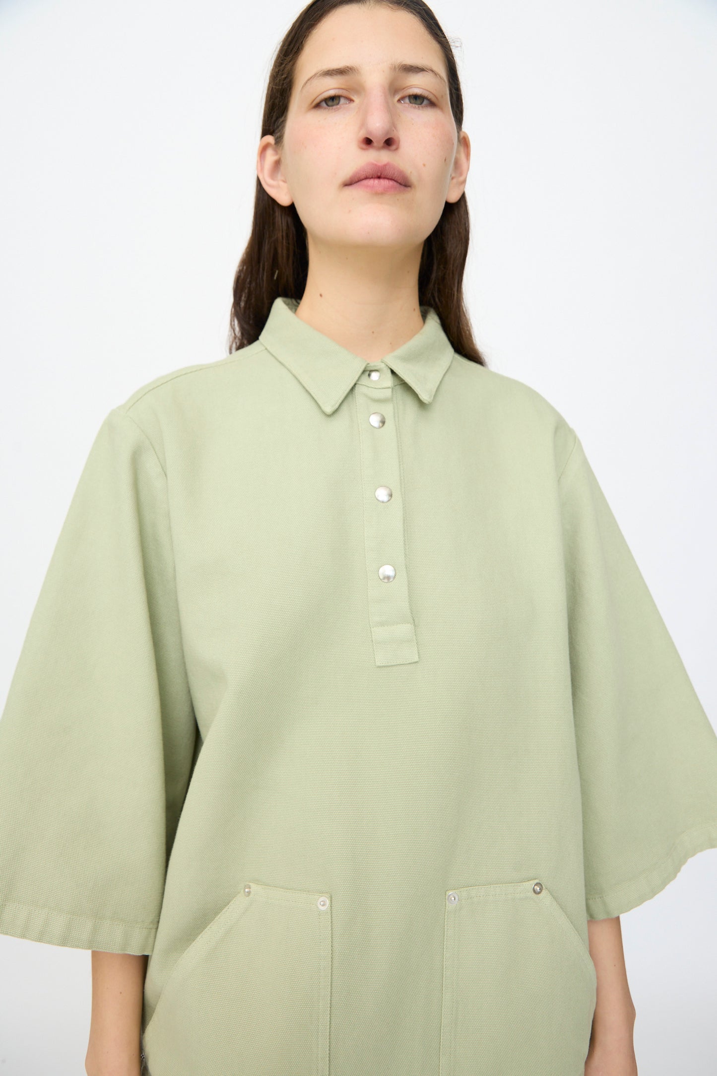 A person with long brown hair wears the AVN Joyeux Dress in Sage, featuring a pale green textured cotton fabric, collared neckline, front buttons, and two pockets, standing against a plain light background.
