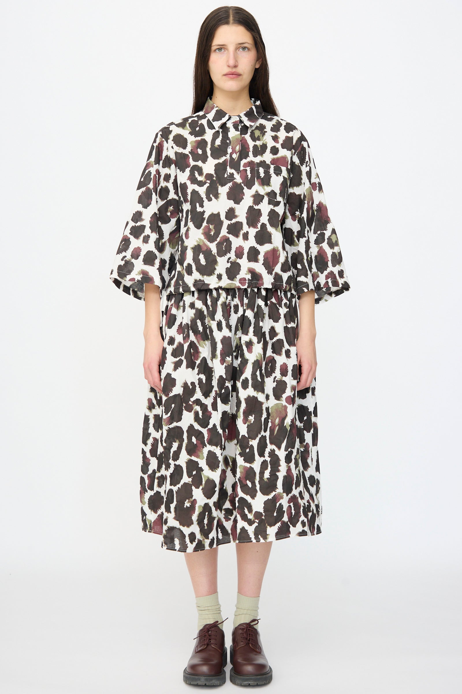 A person stands facing forward wearing the AVN Joyeux Top in White with Print, styled loosely with a matching midi skirt featuring a brown and black animal print, paired with socks and brown shoes.