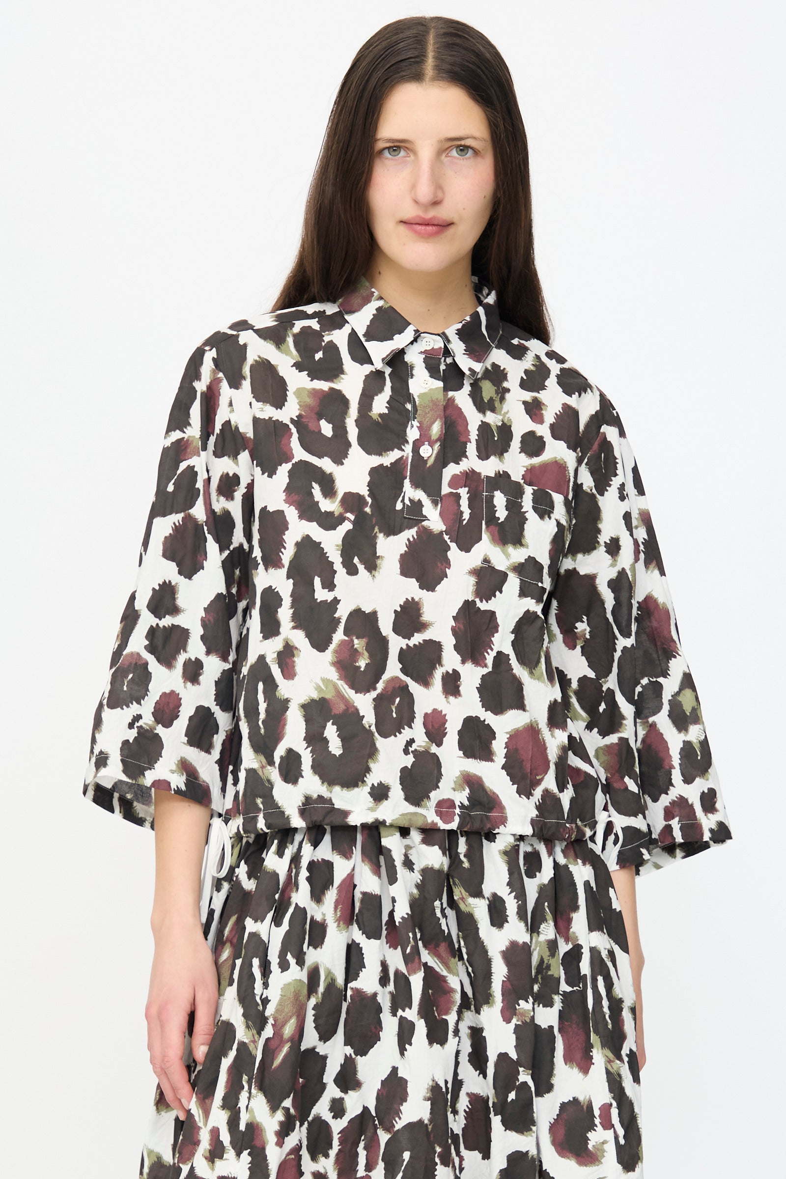 A woman wearing the AVN Joyeux Top in White with Print and a matching pleated skirt stands against a plain white background.