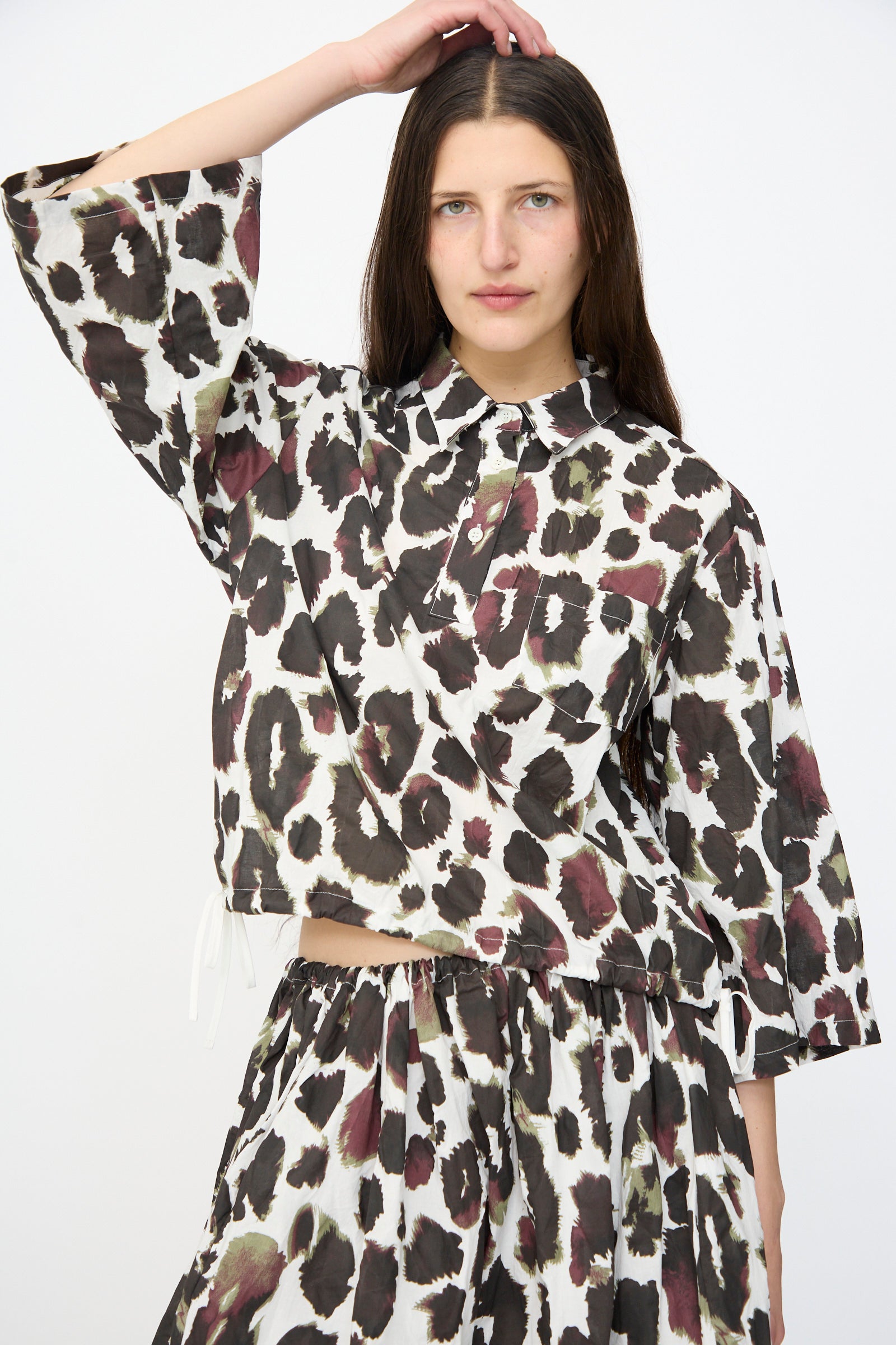 A woman wears the AVN Joyeux Top in White with Print—a loose, 3/4 sleeve cotton blouse—paired with a matching skirt, posing with one hand on her head against a plain white background.