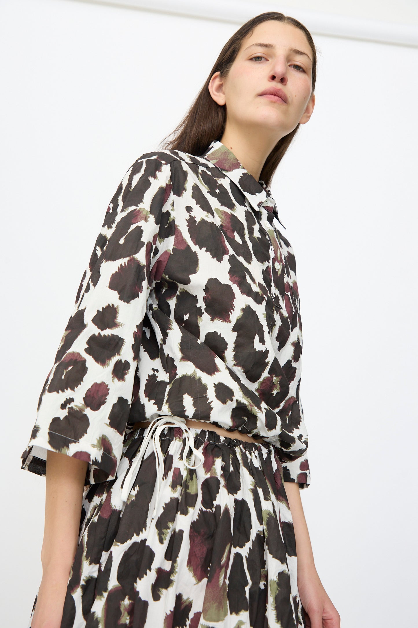 A person wears the AVN Joyeux Top in White with Print, a loose-fitting, 3/4 sleeve cotton top featuring a black and brown abstract spotted pattern, paired with a matching skirt, standing against a plain white background.