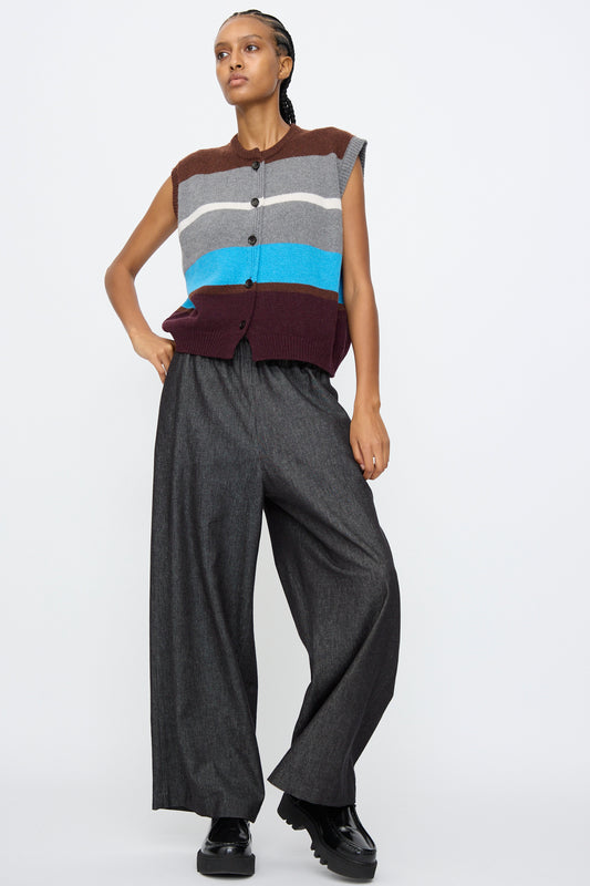 A person stands against a plain white background wearing AVN's Long Easy Pants in Navy, featuring a wide leg and elasticated waistband, paired with a sleeveless striped sweater vest.
