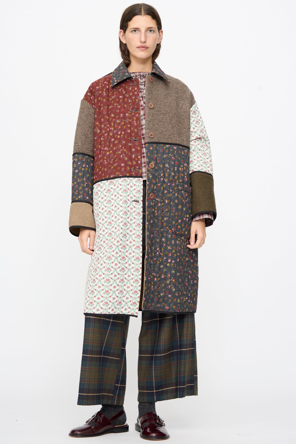 A person wearing the AVN Mid Season Coat in Camel unbuttoned, a knee-length floral patchwork coat paired with plaid trousers and burgundy shoes.