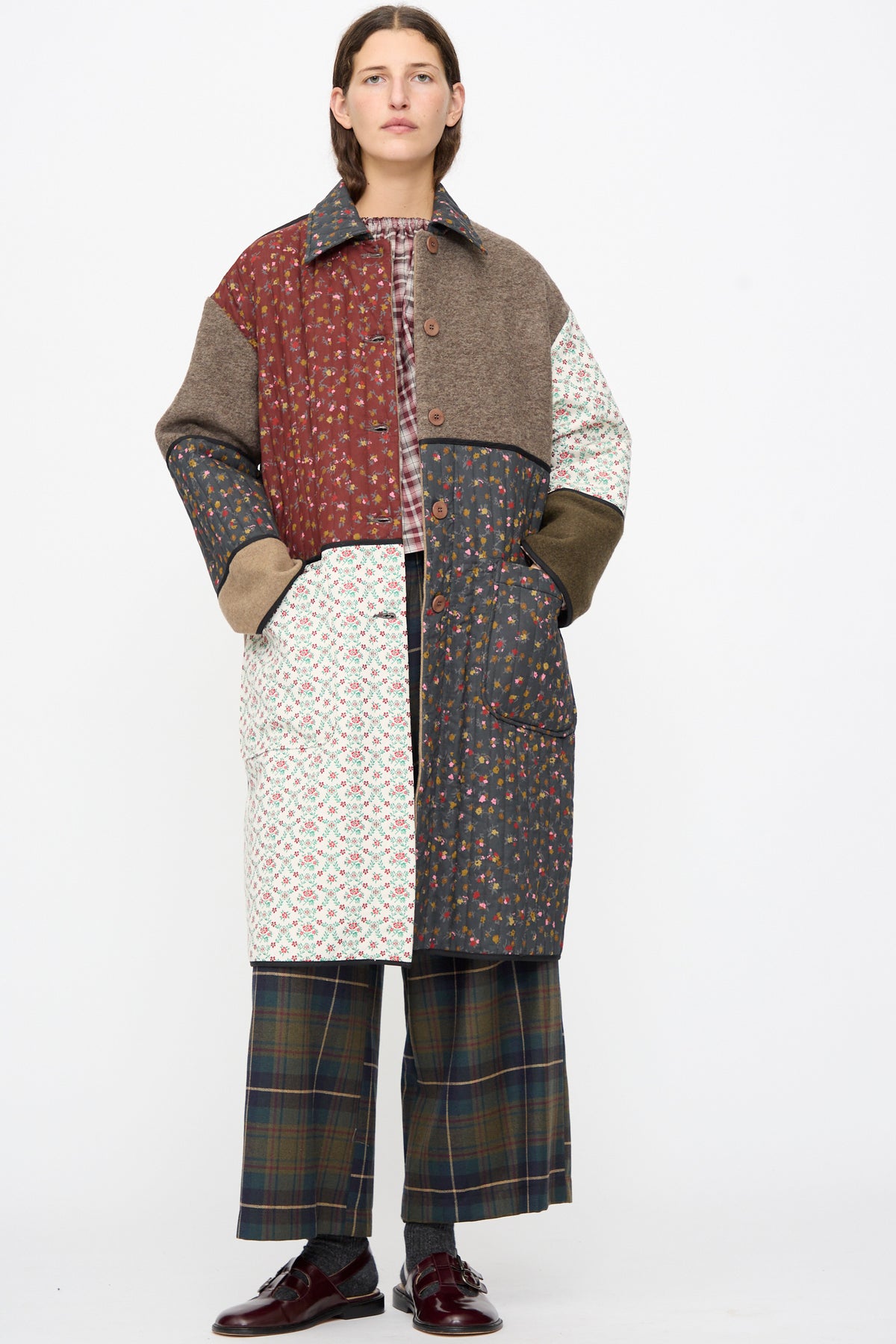 A person wearing an AVN Mid Season Coat in Camel, a knee-length floral patchwork design with solid and quilted panels, paired with plaid wide-leg trousers and dark loafers.