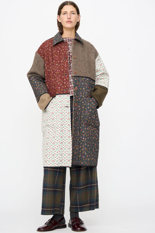A person wearing an AVN Mid Season Coat in Camel, a knee-length floral patchwork design with solid and quilted panels, paired with plaid wide-leg trousers and dark loafers.