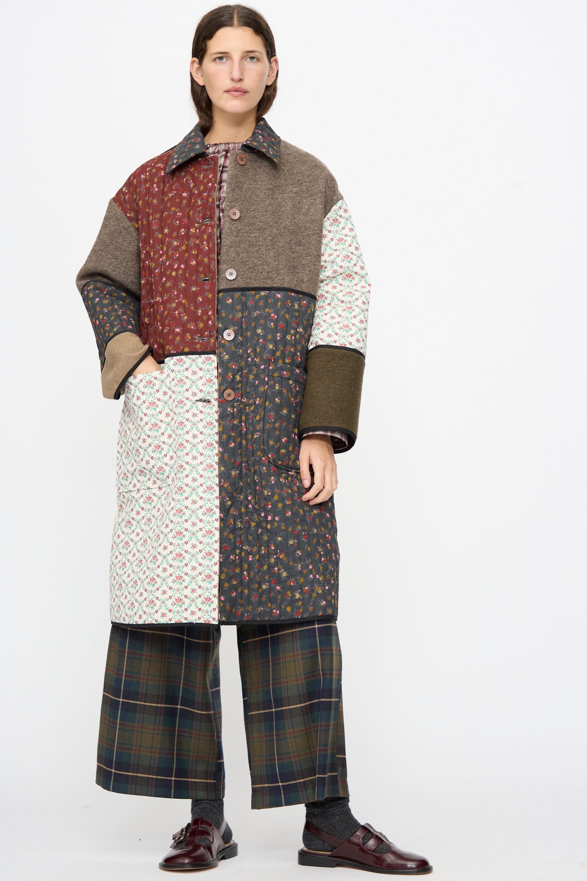 A person wearing an AVN Mid Season Coat in Camel, a long, floral patchwork coat featuring quilted panels, styled with plaid wide-leg trousers and dark loafers.