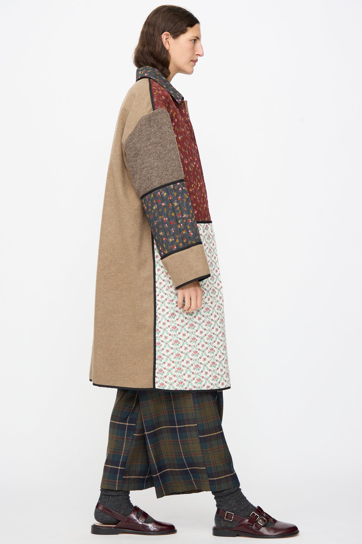 A profile of a person wearing an AVN Mid Season Coat in Camel, a knee-length floral patchwork design, styled with plaid wide-leg trousers and burgundy buckled shoes.
