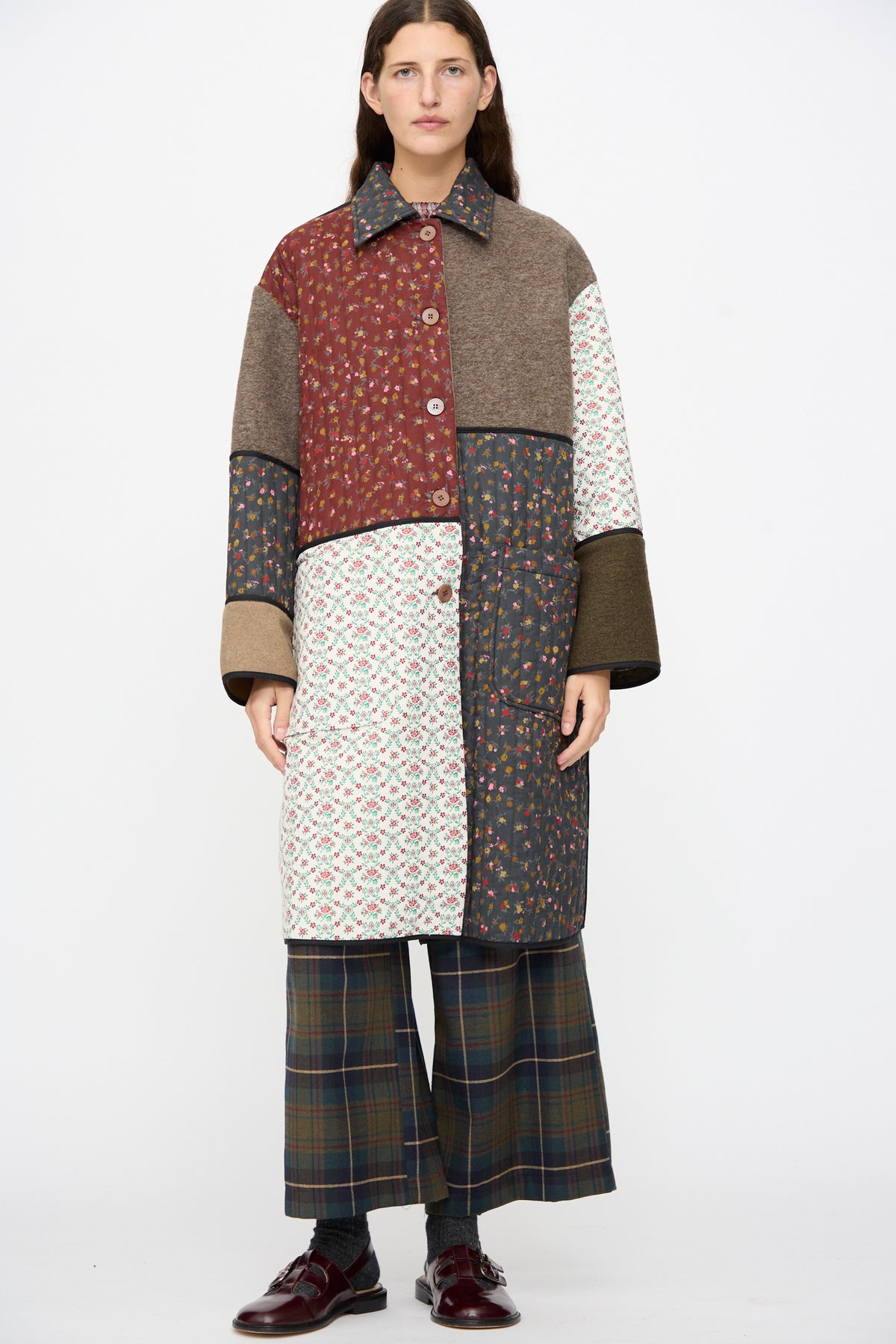 A person wearing the AVN Mid Season Coat in Camel buttoned, a knee-length floral patchwork design with solid and quilted panels, paired with plaid wide-leg trousers and dark loafers.
