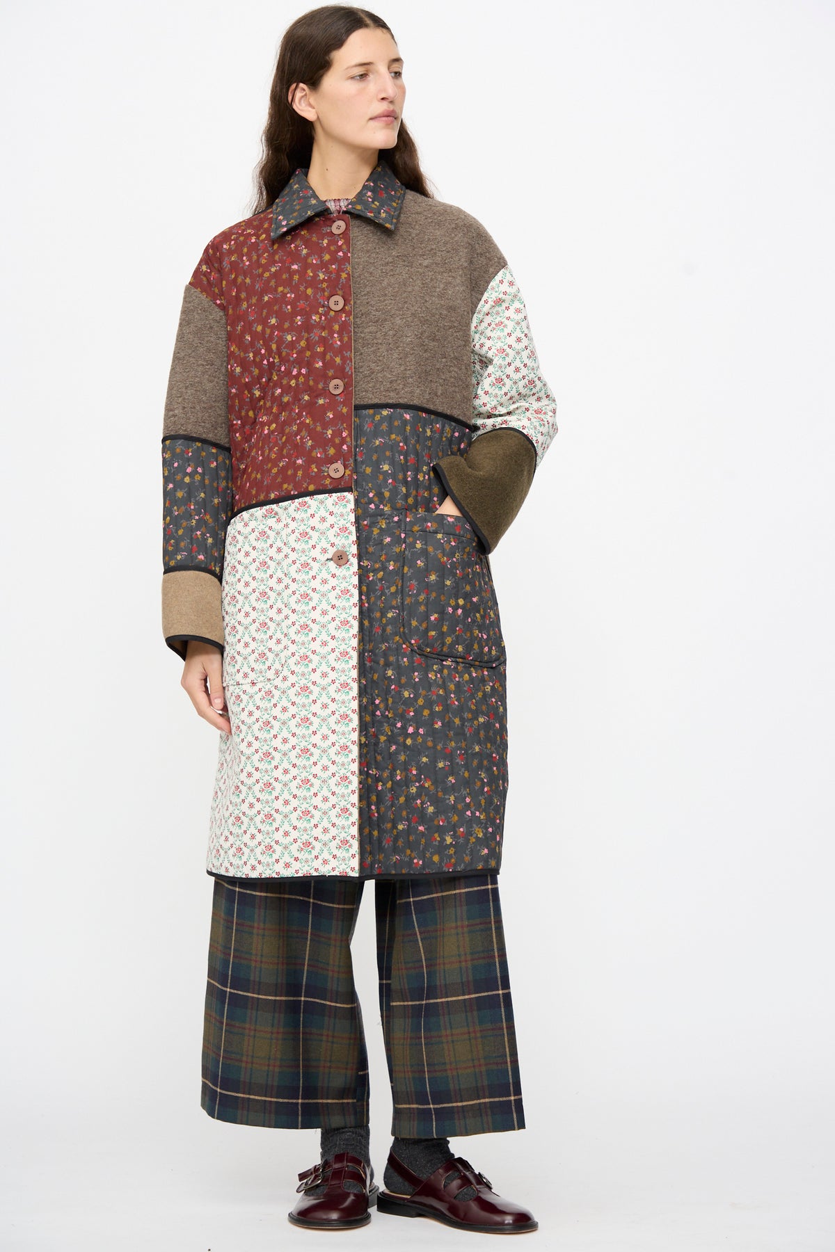 A person wears an AVN Mid Season Coat in Camel, a knee-length floral patchwork wool coat, paired with plaid wide-leg trousers and dark loafers.