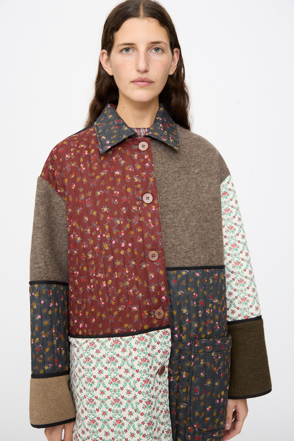 A person wearing an AVN Mid Season Coat in Camel featuring panels of solid brown fabric and various floral patchwork patterns.