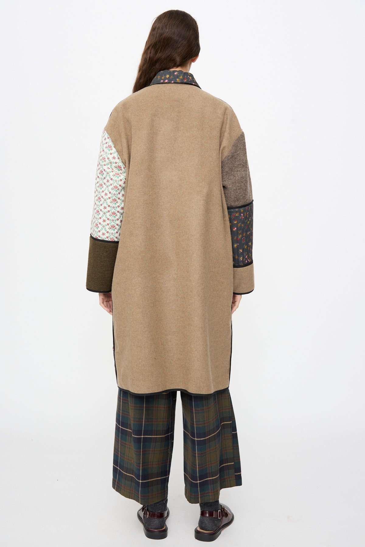 Back view of a person wearing an AVN Mid Season Coat in Camel with floral patchwork sleeves and plaid trousers.