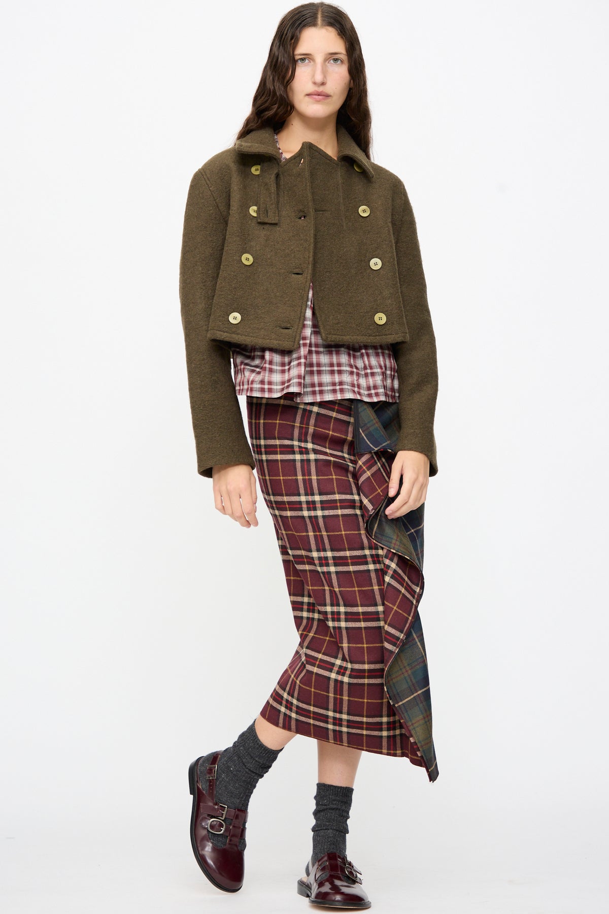 A model wears an AVN Mini Peacoat in Khaki over a plaid shirt and a long, ruffled plaid skirt, paired with grey socks and burgundy buckled shoes.