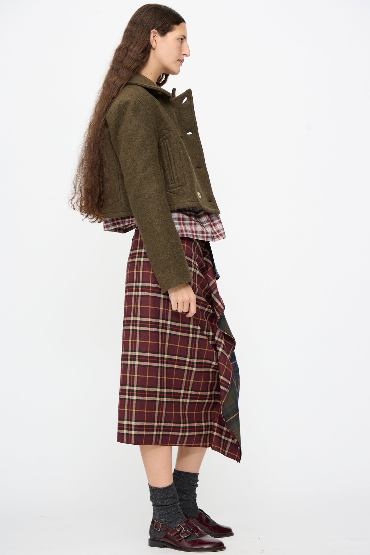 Profile of a person wearing an AVN Mini Peacoat in Khaki over a plaid shirt and ruffled plaid midi skirt, paired with dark double-buckle shoes.