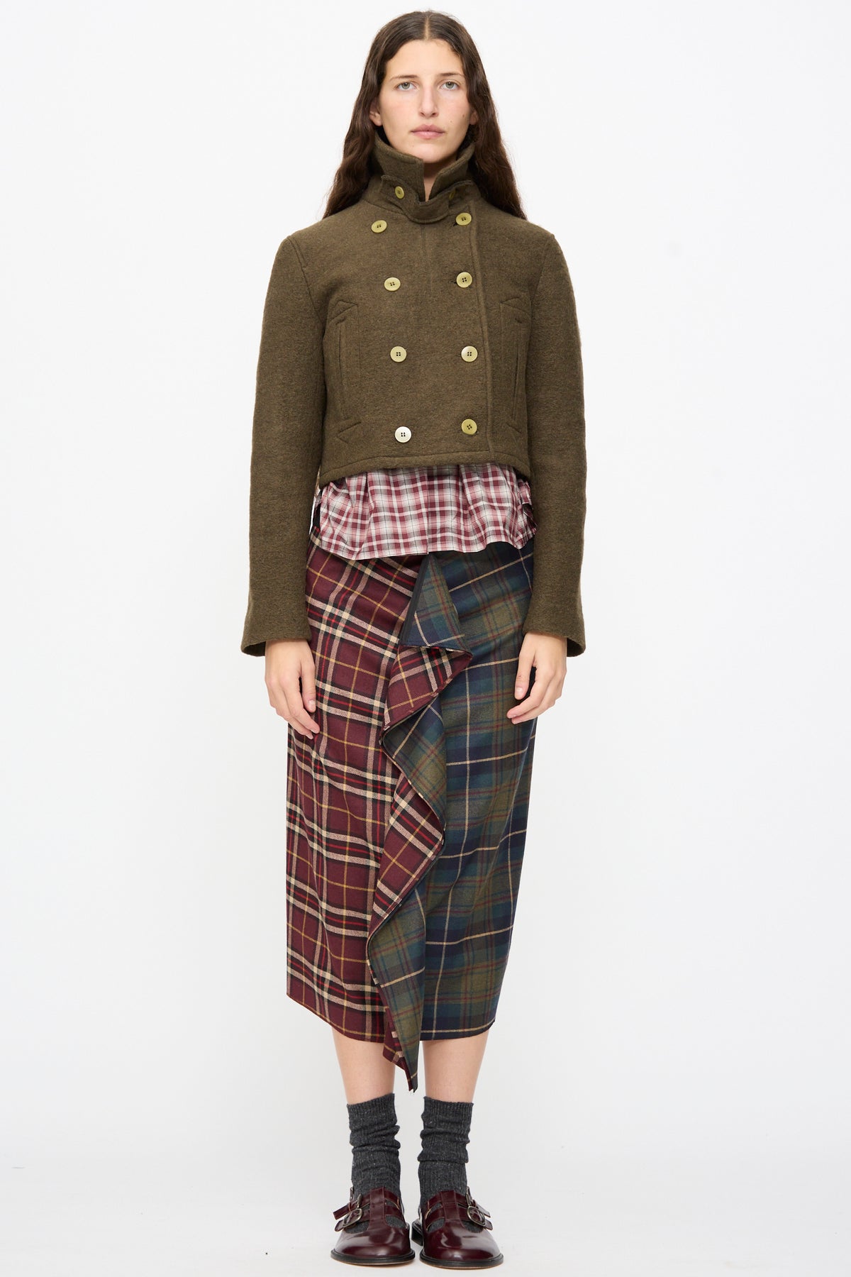 A person wears a cropped, olive Mini Peacoat in Khaki with a high collar over a plaid shirt and contrast red and green plaid skirt.