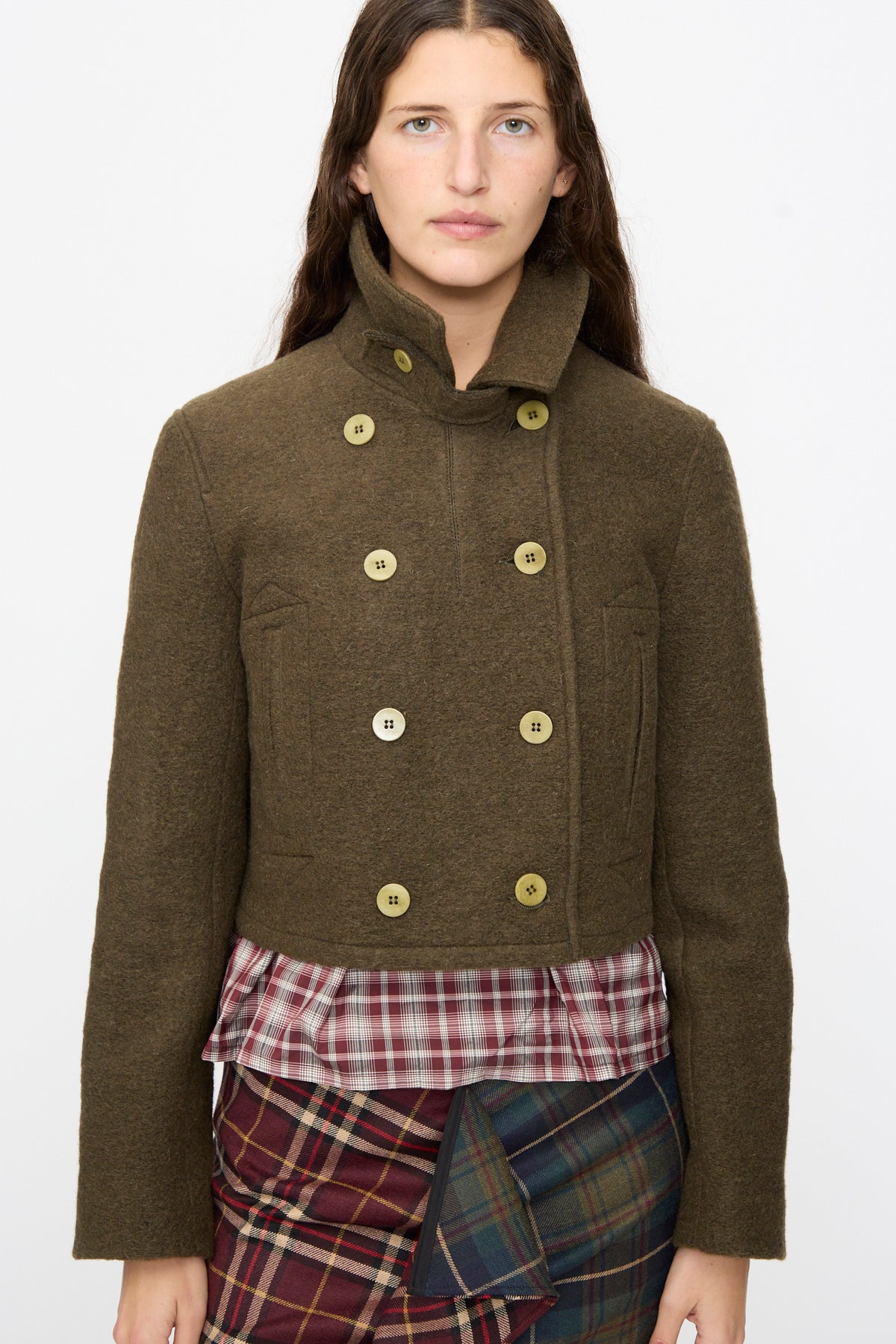 A person wears a cropped, olive Mini Peacoat in Khaki with a high collar over a plaid shirt and contrast red and green plaid skirt.