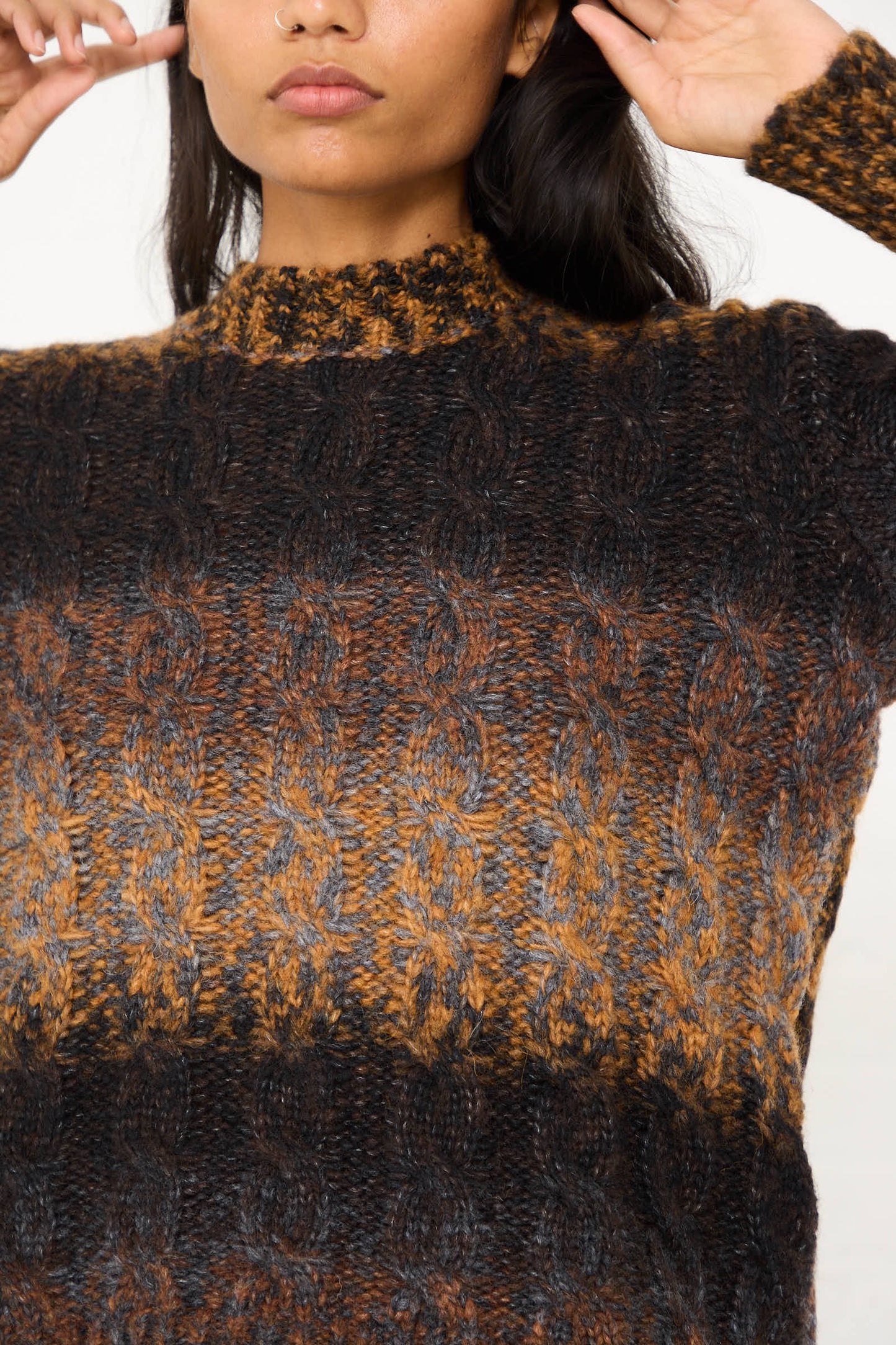 A person wearing the AVN Mountain Sweater in Brown Melange, a relaxed fit cable knit sweater in a multi-color wool blend of brown, black, and grey, shown from the shoulders to the waist, with arms raised and hands near their head.