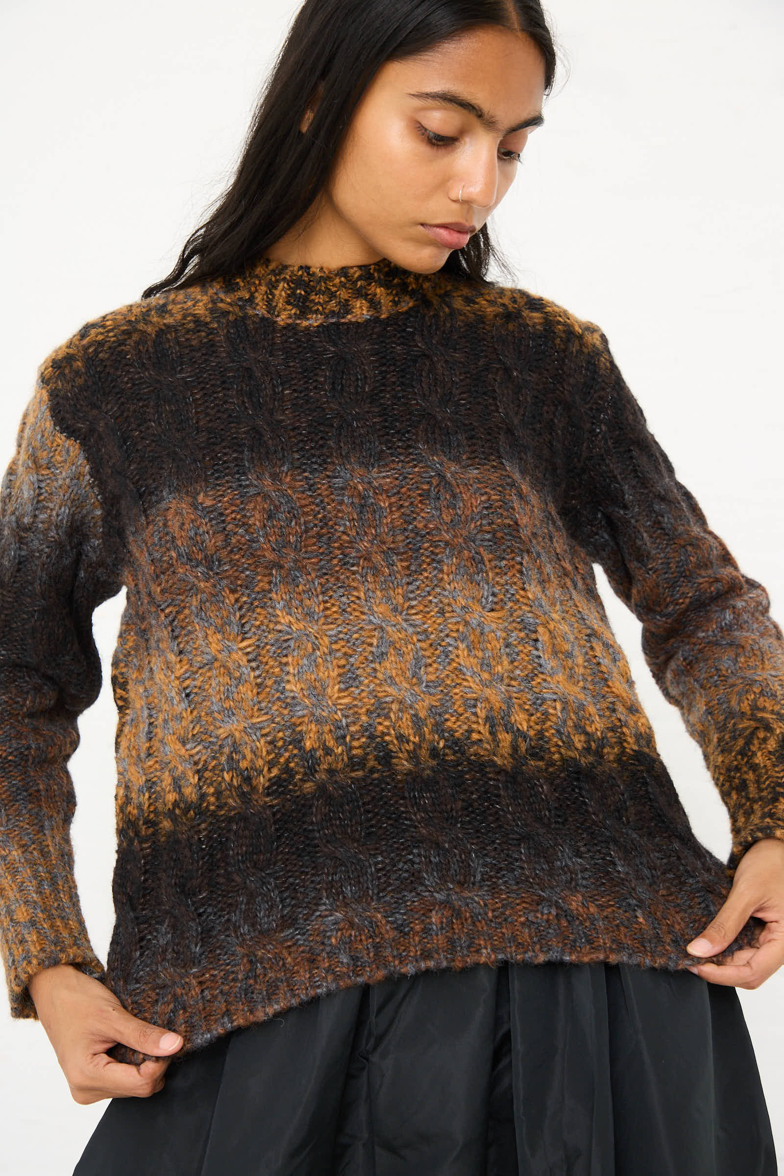 A woman with long dark hair wearing the AVN Mountain Sweater in Brown Melange gazes downward while holding the hem of the relaxed fit garment.