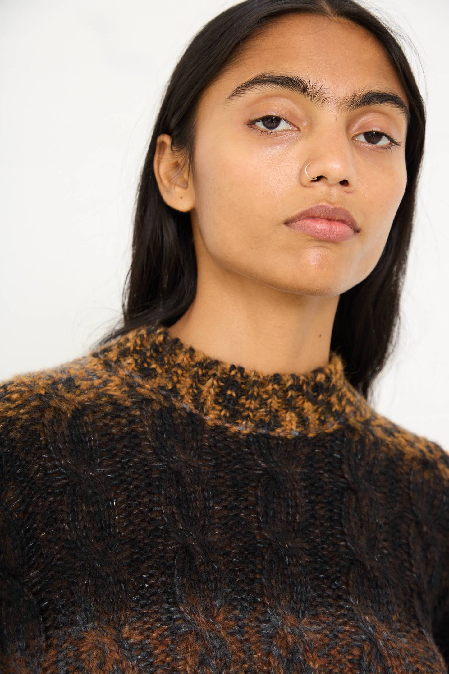 A person with long dark hair and a nose ring wears a relaxed-fit Mountain Sweater in Brown Melange by AVN and looks at the camera.