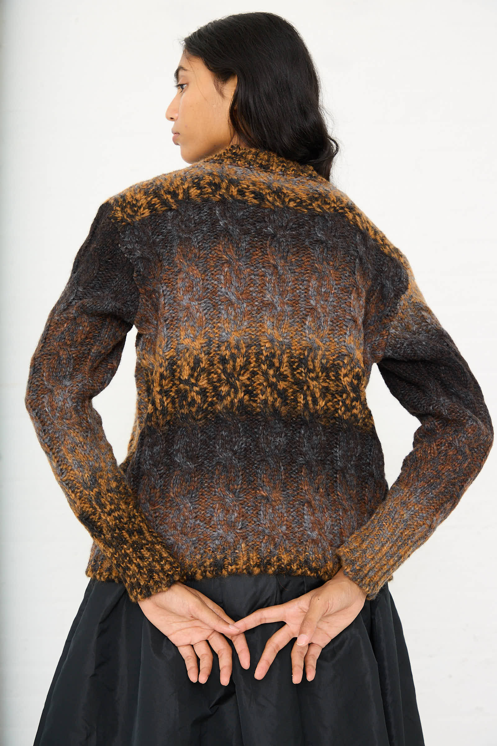 A person with long dark hair stands with their back to the camera, wearing a relaxed fit Mountain Sweater in Brown Melange by AVN and a black skirt, hands clasped behind their back.