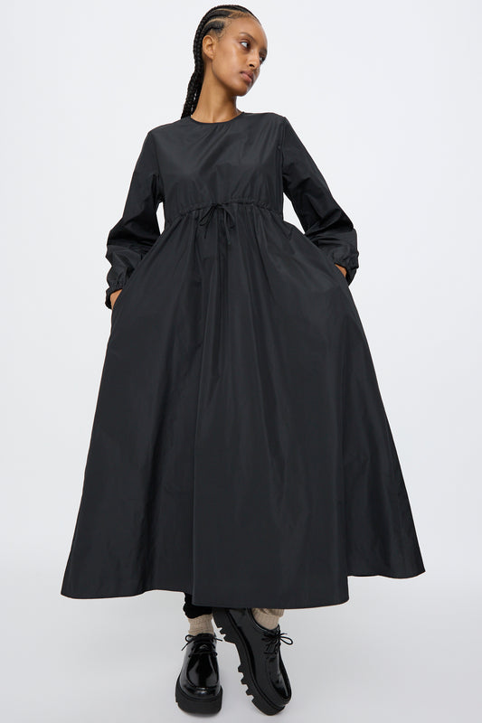 A person stands against a plain white background wearing the AVN New Gem Dress in Black, featuring bishop sleeves and an adjustable tie waist, paired with black shoes and light socks.