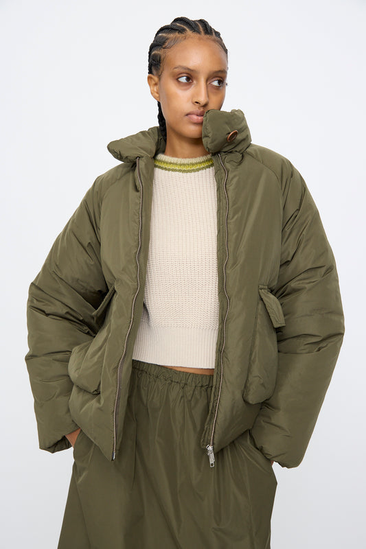 A person wears the AVN Piumino Jacket in Military Green, styled oversized over a beige knit sweater and coordinating olive green skirt, standing against a plain light background.