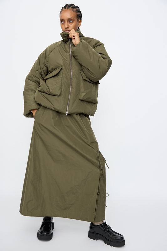 A model wears the AVN Piumino Jacket in Military Green with an oversized fit, styled with a matching long skirt and black platform shoes, standing against a plain white background.