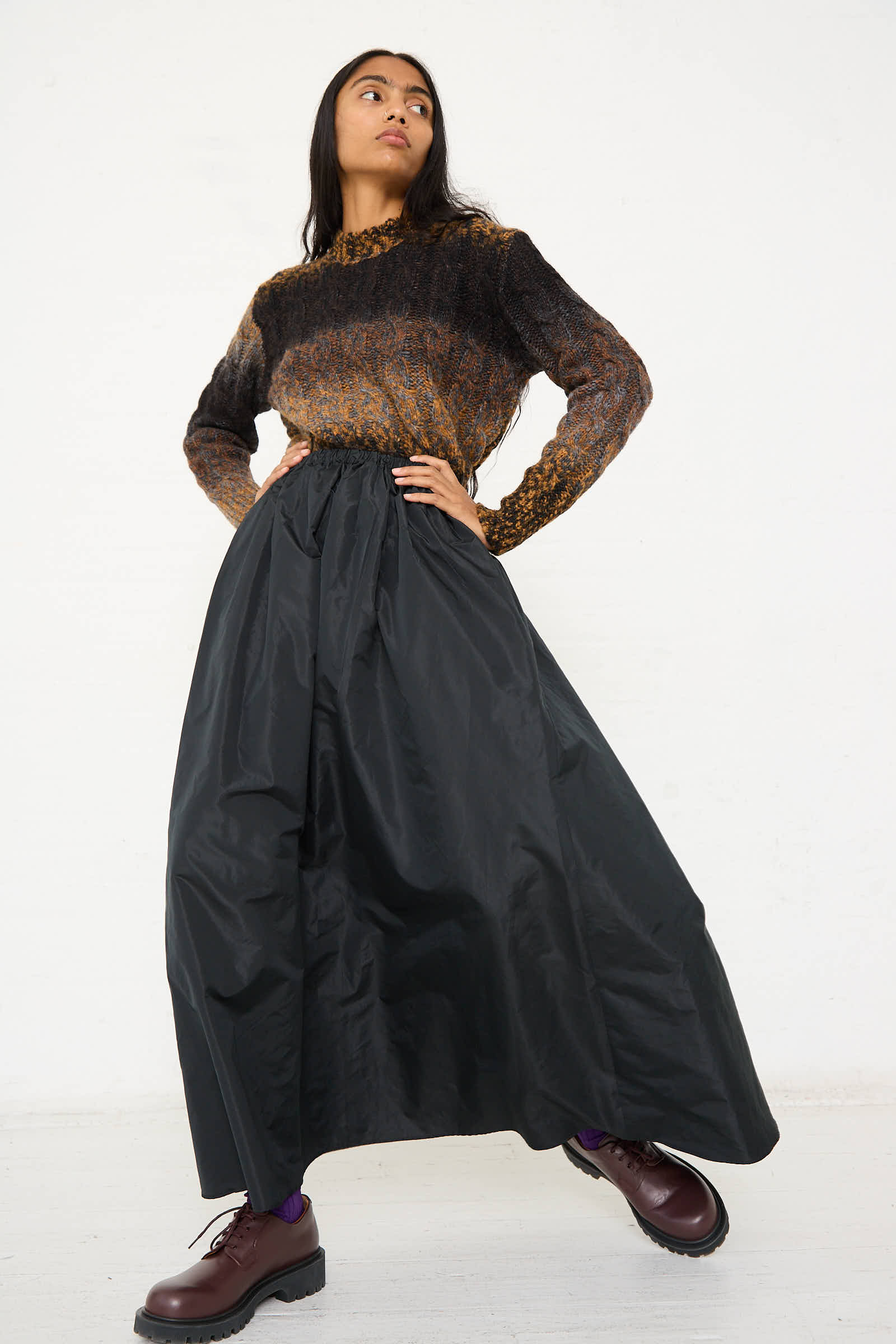 A person stands against a plain background wearing a long-sleeve sweater, the Serenity Skirt in Black from AVN with an A-line silhouette, and dark shoes.