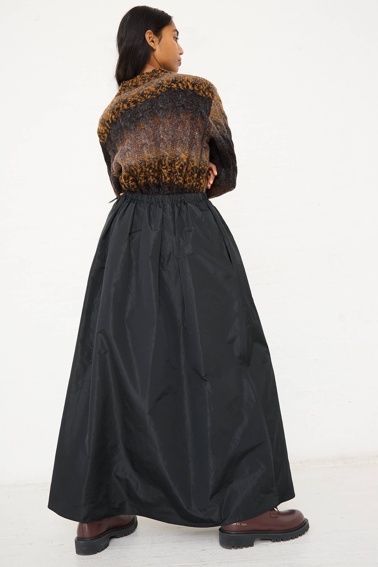 A person with long hair stands facing away, wearing a brownish sweater, dark chunky boots, and an AVN Serenity Skirt in Black. This A-line silhouette black taffeta maxi skirt features pockets and an elasticated waist. The plain white wall serves as the backdrop.