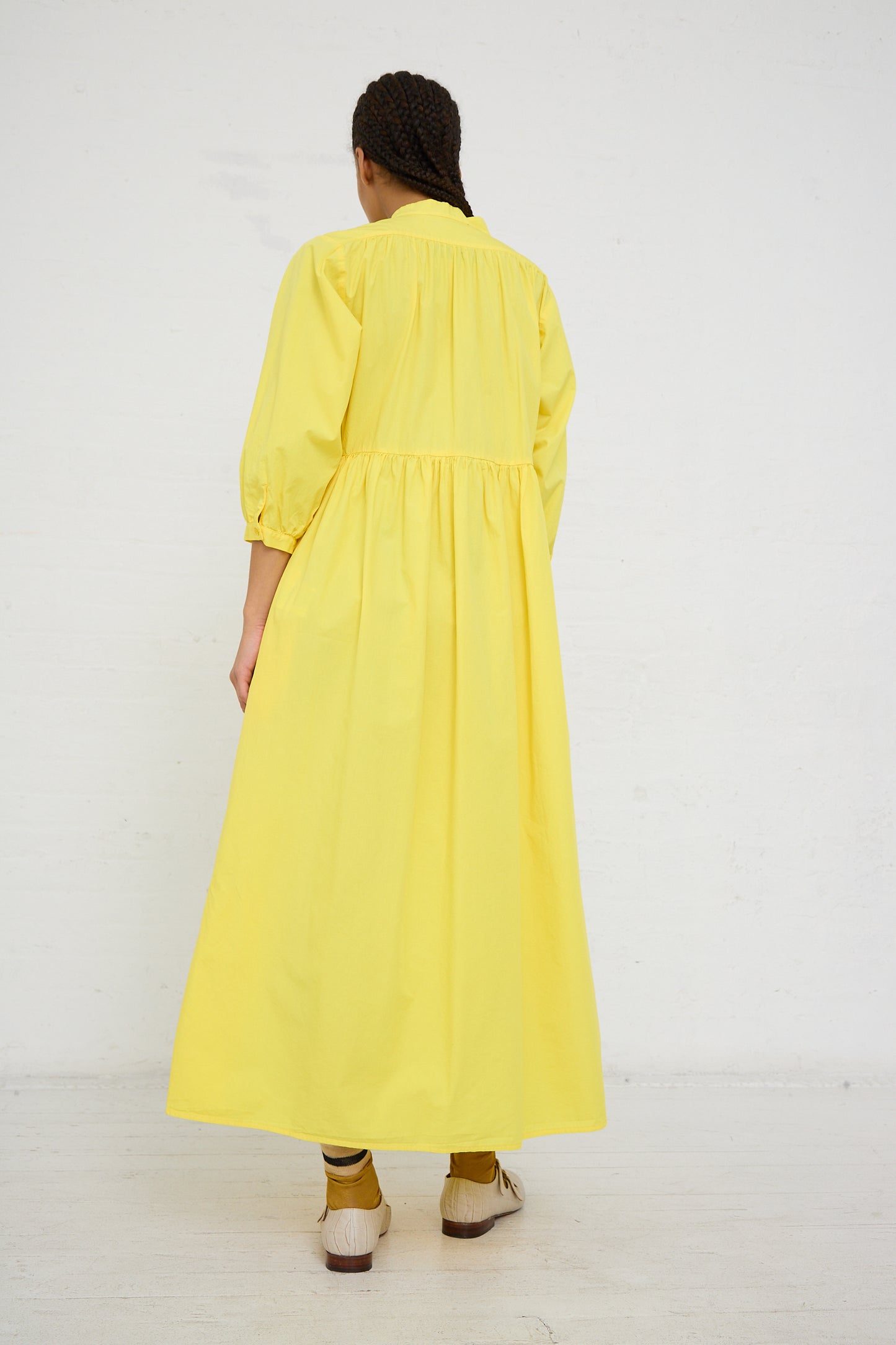 Woman in an AVN Sunshine Dress in Yellow, a long-sleeve, lightweight cotton maxi dress, viewed from behind against a white background.