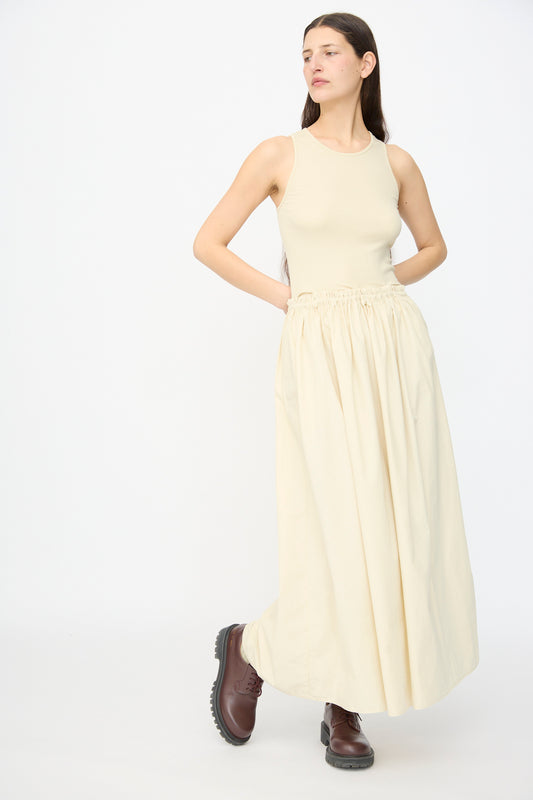 A woman wears the AVN Tank Dress in Sand, a sleeveless beige design paired with dark brown chunky boots, standing against a white background. This elegant look is made in Italy.